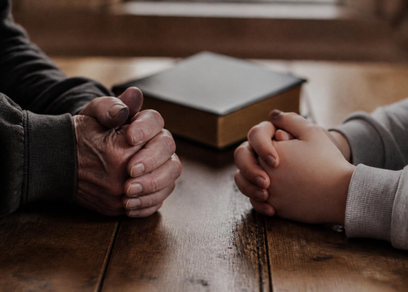 An older hand and a child’s hand folded together in prayer at a wooden table